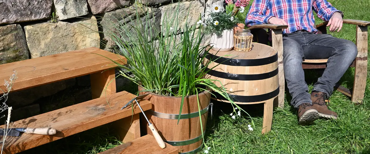 Ein Stimmungsbild im Garten auf dem Man eine Holztreppe, Pflanzkübel Tisch und einen Stuhl sieht. Auf dem Stuhl sitzt eine Person.